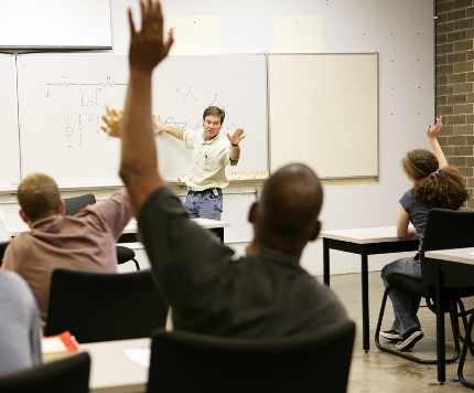 An instructor faces a class of highly engaged students at desks, several with their hands raised.