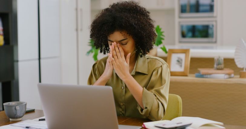 A woman with dark hair sits at a laptop with her hands clasped at her face, showing exhaustion and overwhelm