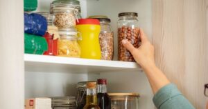 A woman's hand reaches for a jar on a pantry shelf; other staple items are on the shelf.
