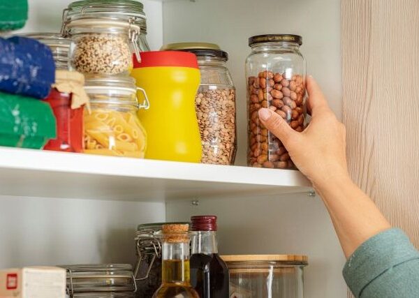 A woman's hand reaches for a jar on a pantry shelf; other staple items are on the shelf.