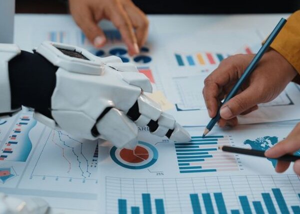 Several hands rest on a page of charts, collaborating on editing. 3 hands are human; one is a robot's.