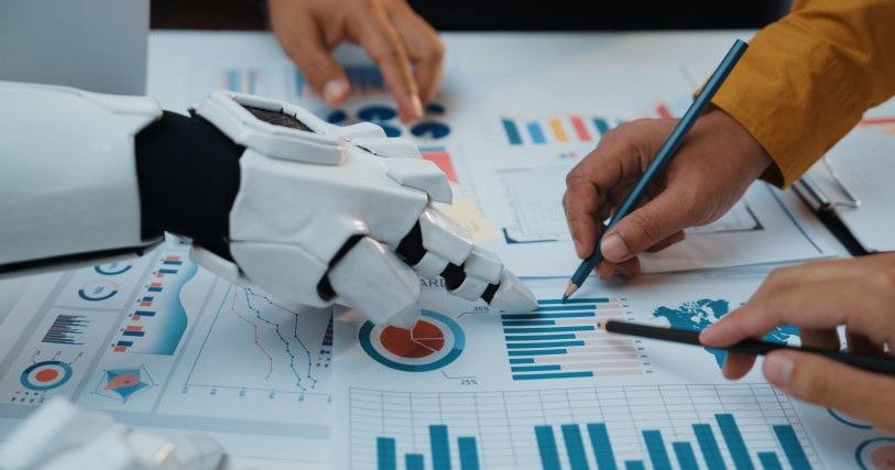 Several hands rest on a page of charts, collaborating on editing. 3 hands are human; one is a robot's.