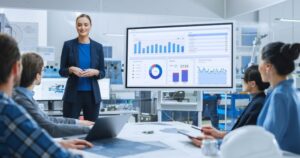 A young woman in a dark blue suit stands next to a large chart and explains to 4 colleagues what the data show.