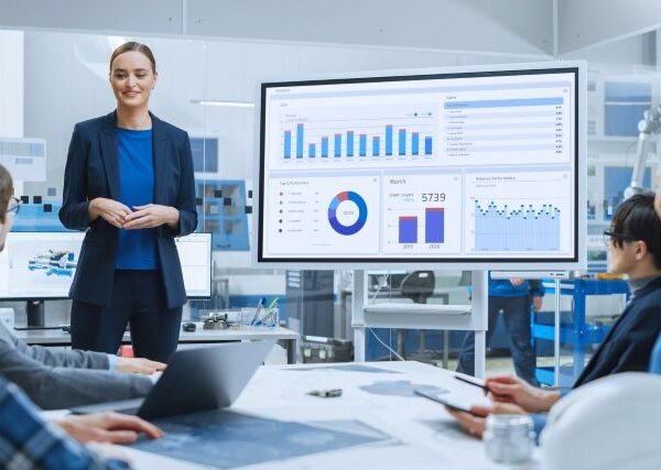 A young woman in a dark blue suit stands next to a large chart and explains to 4 colleagues what the data show.