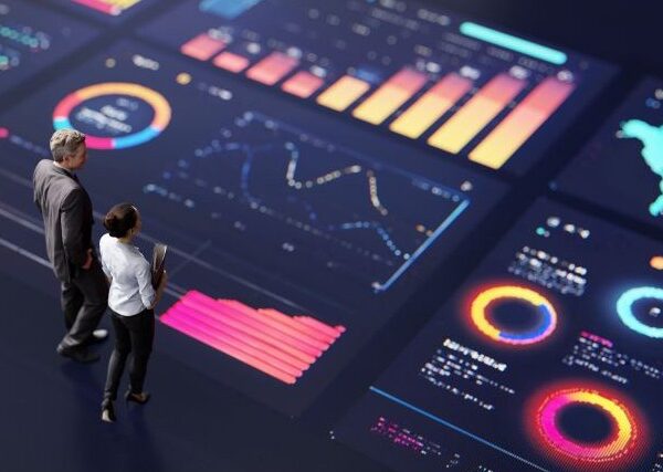 Two small figures, a man and a woman dressed in gray and white business attire, stand on a multicolored display of charts and graphs
