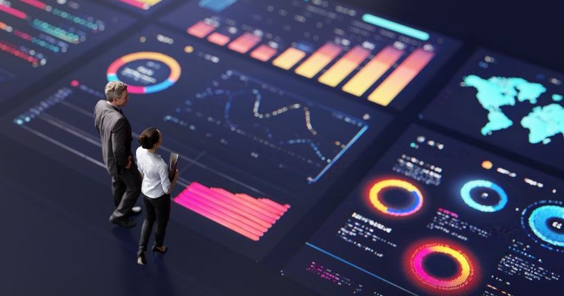 Two small figures, a man and a woman dressed in gray and white business attire, stand on a multicolored display of charts and graphs
