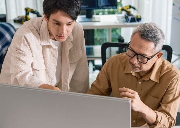 Two Asian men cluster near a computer screen; one watches as the other points to something he is explaining.