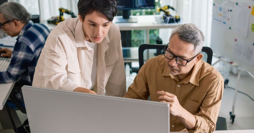 Two Asian men cluster near a computer screen; one watches as the other points to something he is explaining.