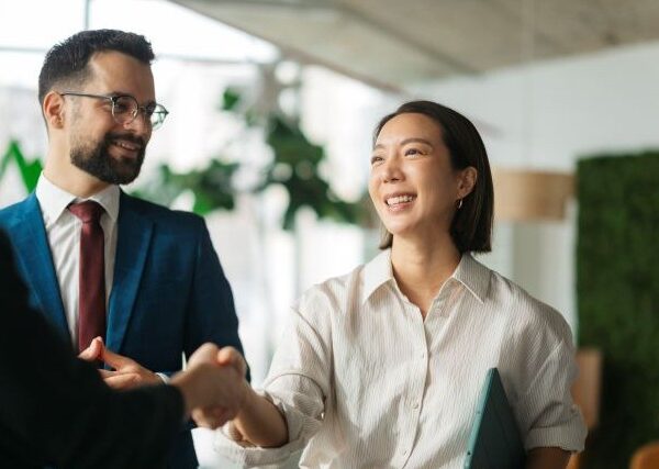 A young Asian woman shakes hands with a colleague while a man in a blue suit jacket looks on.