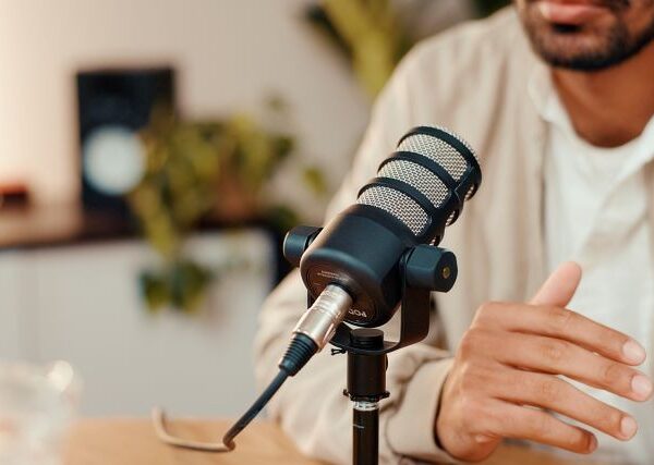 A bearded man, seen from the chin down, speaks into a large black microphone.