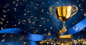 A large gold trophy cup, with a blue ribbon floating around it signifies winning. It stands against a dark background with gold confetti falling around it.