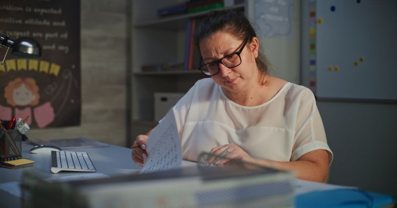 A woman wearing a white shirt and black glasses looks at some papers with a puzzled expression. A computer keyboard is on the table near the papers.