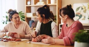Three dark-haired women sit in a row. The one in the center is teaching the others to build a pottery bowl.
