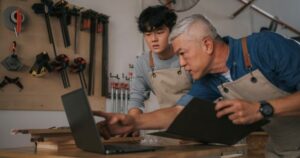 An older man leans over a laptop and instructs a younger man standing next to him They are in a workshop with tools hanging on the wall next to them.