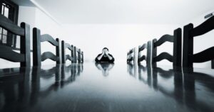A person sits at the head of a long table, with empty chairs along both sides. Their head in in their hands, signaling frustration or overwhelm. The image in in shades of gray.