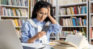A woman with dark hair and glasses sits at a table with a laptop, an open book, and notepads. Packed bookshelves are behind her. She's looking at her phone with a puzzled expression and is holding her head with her left hand.