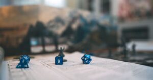 Blue game dice rest on a piece of paper with game instructions with blurry gamepieces in the background