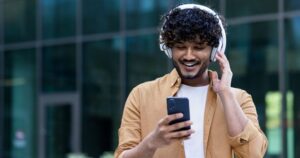 A young man with dark, curly hair and white headphones looks at his smartphone and smiles