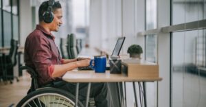 Man seated in a wheelchair, using headphones, smiles and looks attentively at a laptop screen